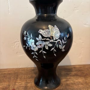 May include: Black vase with a flared rim and base, featuring a decorative design of birds and flowers. The birds and flowers are inlaid with iridescent mother-of-pearl. The vase is a decorative home accent.