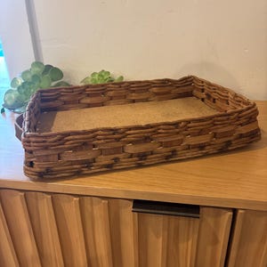 May include: Rectangular woven wicker basket with a cork bottom. The basket is brown and has a rectangular shape. The basket is sitting on a wooden surface.
