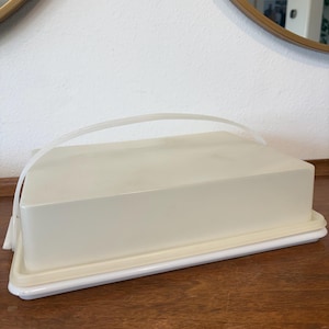 May include: An off-white plastic cake carrier with a clear handle. The rectangular carrier has a white base and lid. The carrier is on a wooden surface.