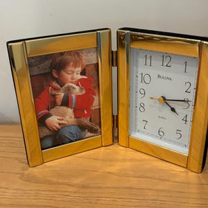 Puede incluir: Una combinación de marco de fotos y reloj con bisagras, de color dorado. El lado izquierdo muestra una fotografía, mientras que el lado derecho presenta un reloj con la marca "Bulova". El reloj tiene una esfera blanca con números y manecillas negras.