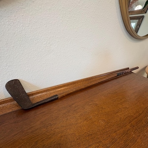 Könnte beinhalten: Ein antiker Golfschläger mit einem dunklen Holzschaft und einem dunklen Metallschlägerkopf. Der Schläger liegt auf einer dunklen Holzoberfläche, mit einer weißen Wand im Hintergrund. Ein goldgerahmter Spiegel ist im Hintergrund zu sehen.