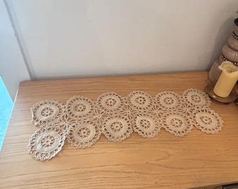 May include: A cream-colored, crocheted lace table runner with a repeating circular floral design. The runner is displayed on a light wood surface, showcasing its delicate openwork pattern. The runner is approximately 12 inches wide and 36 inches long.
