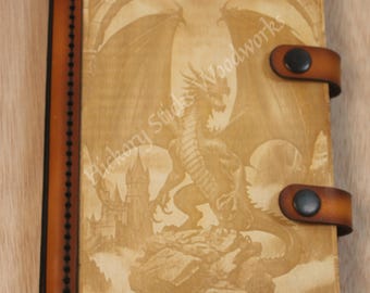 May include: A wooden journal with a detailed dragon engraving on the cover. The journal features a leather spine and closure straps with black button snaps. The text "Hickory Sticks Woodworks" is visible on the cover.