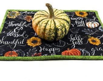 May include: A rectangular quilted fabric mat with a black background featuring sunflowers, orange pumpkins, and white cursive text including "thankful," "blessed," and "Happy Fall Y'all." The mat has a bright green border and a small striped pumpkin.