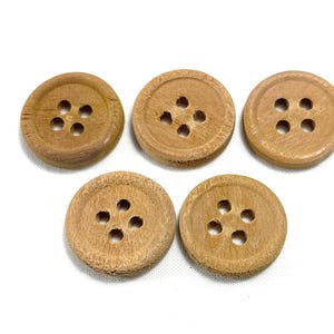 May include: Five round, light brown wooden buttons arranged in a group. Each button features a smooth, circular surface with a slightly raised outer rim and four evenly spaced holes in the center for threading, showcasing a natural wood grain texture.