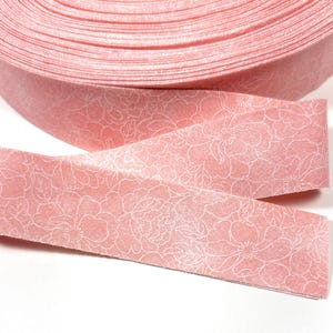 May include: Pink fabric ribbon with a white floral pattern. The ribbon is rolled up and a piece is laying on top of the roll.