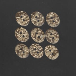 May include: Nine oval-shaped buttons with a speckled, marbled design in cream and brown. Each button has two holes for sewing. The buttons are arranged in a grid pattern against a dark background.