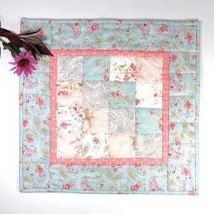 May include: Square quilted placemat with a patchwork design. The center features a grid of floral and patterned squares in shades of pink, gray, and white. A coral border frames the patchwork, set against a light blue floral paisley background.