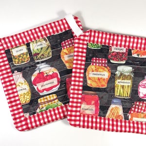 May include: Two square pot holders featuring a red and white gingham border and a central print of glass jars filled with various preserved vegetables and fruits, including labels for olives, okra, red peppers, pineapples, asparagus, tomatoes, and radishes.