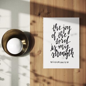 May include: A white print with the text "the joy of the lord is my strength" in black calligraphy. The print is on a wooden surface, next to a brass bowl with a white sphere inside.