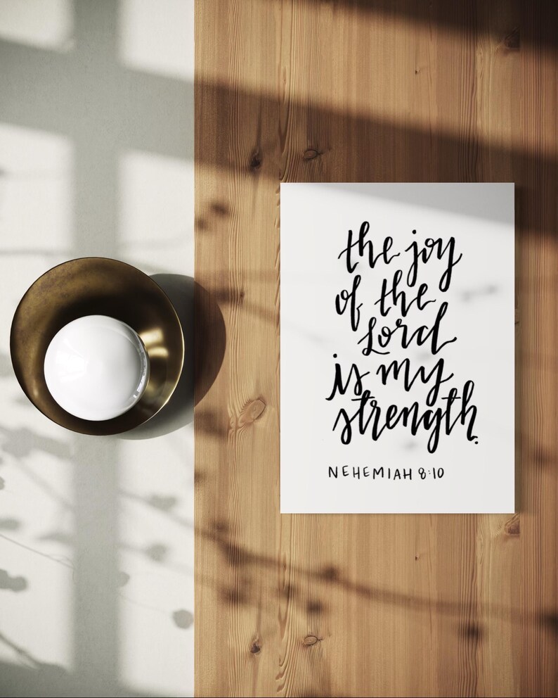 May include: A white print with the text "the joy of the lord is my strength" in black calligraphy. The print is on a wooden surface, next to a brass bowl with a white sphere inside.