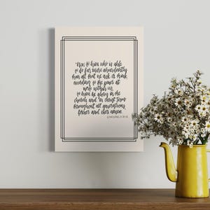 May include: A framed print with a black border features a handwritten quote from Ephesians 3:20-21. The print is on a wooden surface, beside a yellow jug filled with white daisies. The quote is in black script.