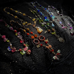 May include: A collection of five necklaces featuring gold-toned chains adorned with various colorful beads, including floral shapes, small rings, and charms like a butterfly, a key, and a teardrop, arranged in a fan pattern on a black fabric.