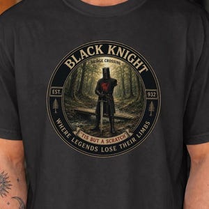 May include: A black t-shirt featuring a circular graphic of a knight in armor standing on a wooden bridge in a forest. The text reads BLACK KNIGHT, BRIDGE CROSSING, EST. 932, and 'TIS BUT A SCRATCH' WHERE LEGENDS LOSE THEIR LIMBS in a circular border.