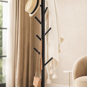 May include: A black coat rack with multiple angled arms, holding a straw hat with a black band, a tan purse, and a cream-colored blanket with tassels. The rack has a tripod base and is in a room with neutral tones.