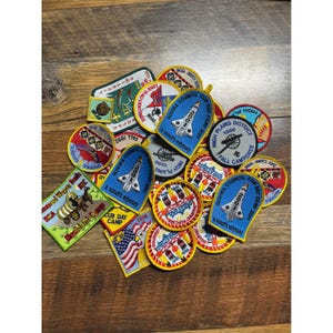Op de afbeelding: Een verzameling kleurrijke geborduurde patches, met verschillende ontwerpen en tekst. De patches bevatten afbeeldingen van raketten, kanonnen en tekst zoals "Cub Day Camp" en "Quivira Council". De patches zijn in verschillende vormen en maten.