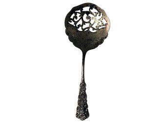 Puede incluir: Una cuchara de servicio de metal plateado con un cuenco ancho y redondo que presenta un intrincado patrón de filigrana calada y detalles de hojas grabadas. El mango está adornado con volutas en relieve y elementos decorativos.