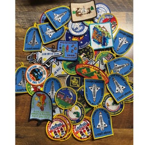 May include: A collection of colorful embroidered patches, featuring various designs and text. The patches include images of rockets, trains, and cartoon characters, with text such as "Fun with Son" and "Cub Day Camp 2001".