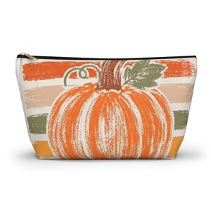 May include: A zippered pouch featuring a central orange pumpkin illustration with a brown stem and green leaf. The background consists of horizontal, distressed-style stripes in shades of orange, beige, olive green, and yellow, creating a textured look.