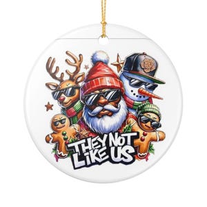 May include: A round Christmas ornament featuring a festive design. The ornament showcases Santa Claus, a reindeer, a snowman, and gingerbread figures, all wearing sunglasses. The text "THEY NOT LIKE US" is displayed in a bold font. A gold string is attached for hanging.
