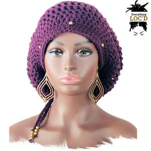May include: A purple crochet hat with gold beads and buttons. The hat has a long tail that can be tied or left loose.