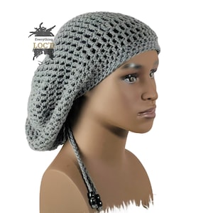 Peut inclure: Béret en crochet gris avec une longue attache nouée. Le béret a un design en maille et est porté sur une tête de mannequin.