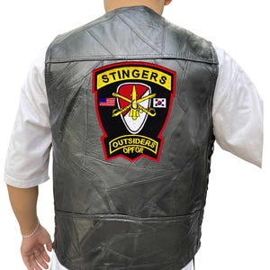 K&ouml;nnte beinhalten: Eine schwarze Lederweste mit einem gen&auml;hten Patchwork-Design. Die Weste hat einen gro&szlig;en Aufn&auml;her auf dem R&uuml;cken mit den Worten "STINGERS" und "OUTSIDERS OPFOR" in gelben Buchstaben. Der Aufn&auml;her enth&auml;lt auch ein Design mit gekreuzten Schwertern und Flaggen.