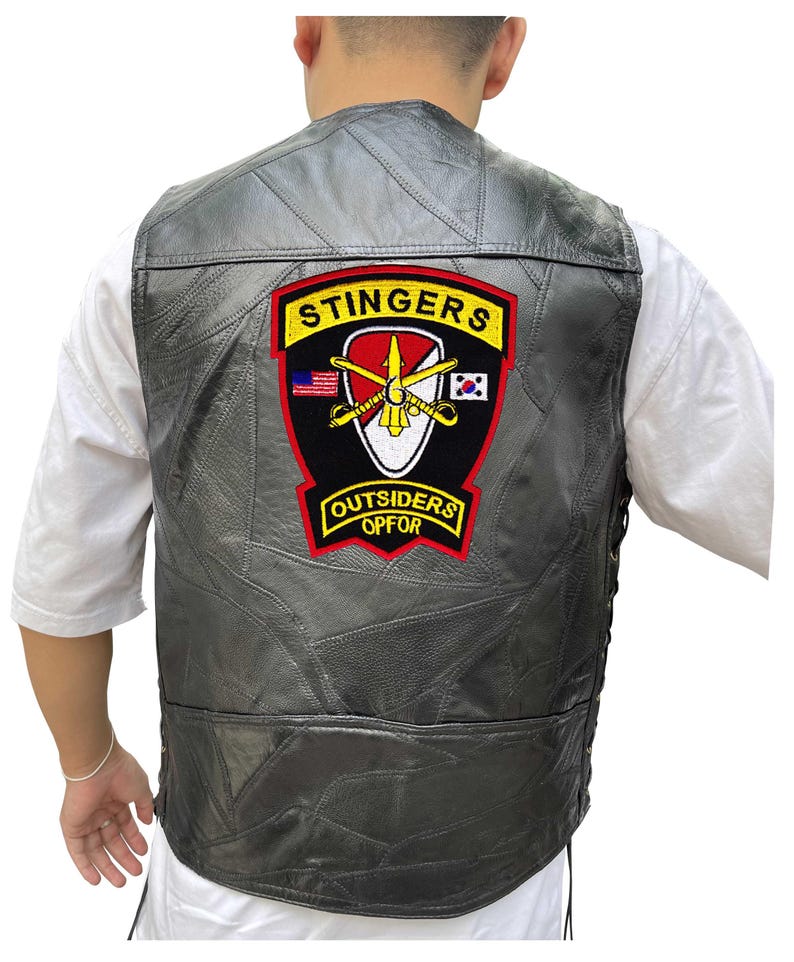 K&ouml;nnte beinhalten: Eine schwarze Lederweste mit einem gen&auml;hten Patchwork-Design. Die Weste hat einen gro&szlig;en Aufn&auml;her auf dem R&uuml;cken mit den Worten "STINGERS" und "OUTSIDERS OPFOR" in gelben Buchstaben. Der Aufn&auml;her enth&auml;lt auch ein Design mit gekreuzten Schwertern und Flaggen.