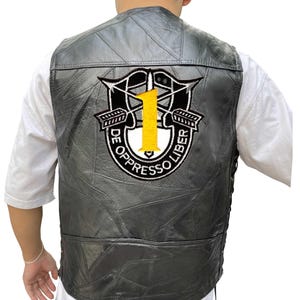 K&ouml;nnte beinhalten: Schwarze Lederweste mit einem aufgen&auml;hten Aufn&auml;her mit einer gelben Zahl 1, gekreuzten Pfeilen und dem Text "DE OPPRESSO LIBER". Die Weste hat ein Patchwork-Design und eine seitliche Schn&uuml;rung.