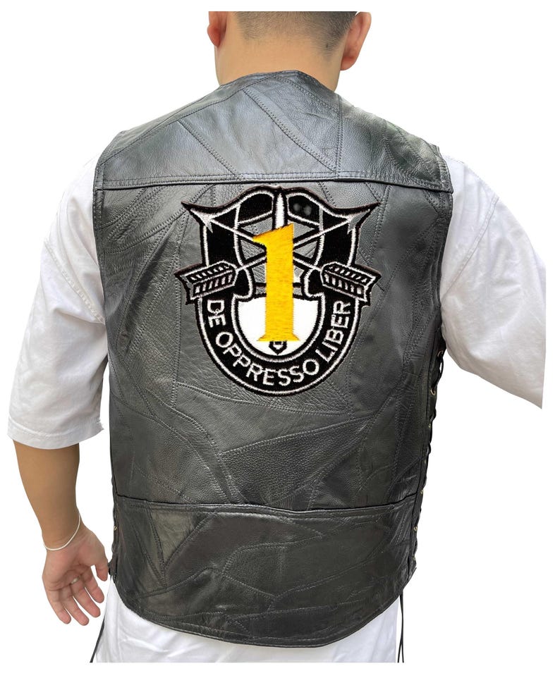K&ouml;nnte beinhalten: Schwarze Lederweste mit einem aufgen&auml;hten Aufn&auml;her mit einer gelben Zahl 1, gekreuzten Pfeilen und dem Text "DE OPPRESSO LIBER". Die Weste hat ein Patchwork-Design und eine seitliche Schn&uuml;rung.