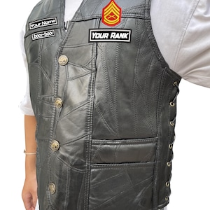 K&ouml;nnte beinhalten: Schwarze Lederweste mit Patchwork-Design, goldenen Kn&ouml;pfen und seitlicher Schn&uuml;rung. Die Weste hat zwei Aufn&auml;her mit der Aufschrift "Your Name" und "Your Rank" sowie ein rot-goldenes Emblem.