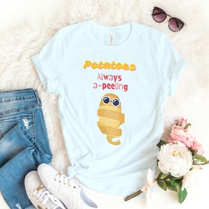 May include: Light blue t-shirt with the text "Potatoes Always a-peeling" and a cartoon potato character wearing sunglasses. The shirt is styled with blue jeans, white sneakers, and sunglasses.