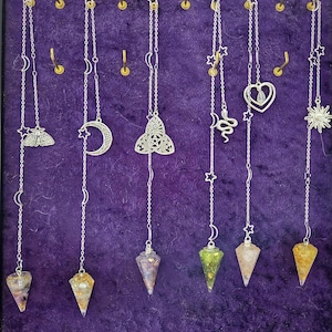 May include: Six silver-toned metal chains hanging from hooks, each featuring celestial charms like stars, moons, a moth, a snake, a heart, and a sun. Each chain terminates in a pointed, cone-shaped crystal or resin pendant with varied internal colors.