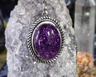 May include: A silver chain necklace with a purple glitter pendant. The pendant is set in a silver oval frame with a rope-like border.