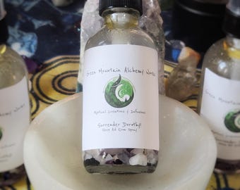 May include: A clear glass bottle with a black spray top filled with a clear liquid and small crystals. The bottle label reads "Green Mountain Alchemy Works", "Mystical Creations & Infusions", "Surrender Dorothy!" and "Sleep Aid Room Spray".