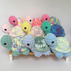 May include: A collection of colorful, crocheted turtle plushies. Each turtle features a textured shell in various pastel shades, including pink, blue, yellow, and green, with contrasting heads and black button eyes. The turtles are displayed on a small, light-colored shelf.