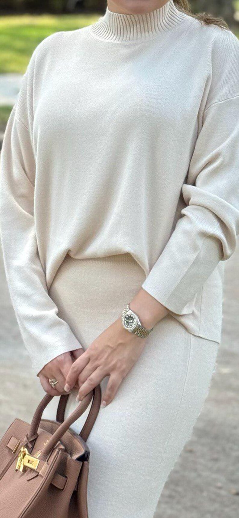 May include: A woman wearing a cream-colored turtleneck sweater and matching skirt. She is holding a brown leather handbag with a gold clasp and wearing a silver watch on her wrist.