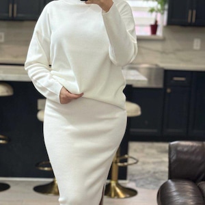 May include: A white turtleneck sweater and matching skirt set. The sweater has long sleeves and a relaxed fit. The skirt is a midi length and has a slit on the side.