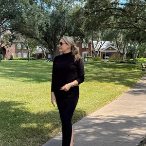 May include: A woman wearing a black mock neck shirt and a black skirt with a slit on the side. She is wearing white sneakers and sunglasses. She is standing on a sidewalk in front of a grassy area.