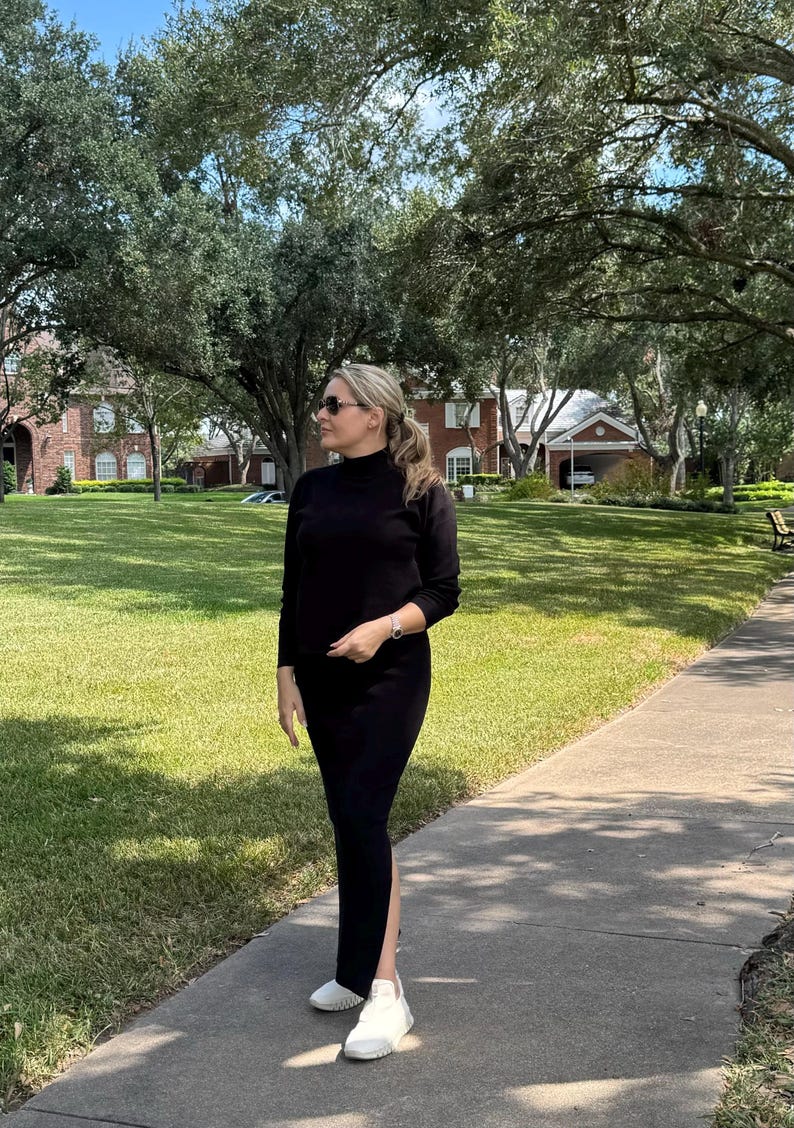 May include: A woman wearing a black mock neck shirt and a black skirt with a slit on the side. She is wearing white sneakers and sunglasses. She is standing on a sidewalk in front of a grassy area.