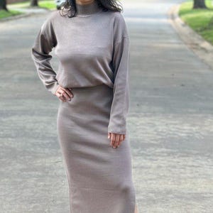 May include: A woman wearing a long-sleeved, light brown sweater dress with a mock neck and a side slit. She is wearing white sneakers and sunglasses.