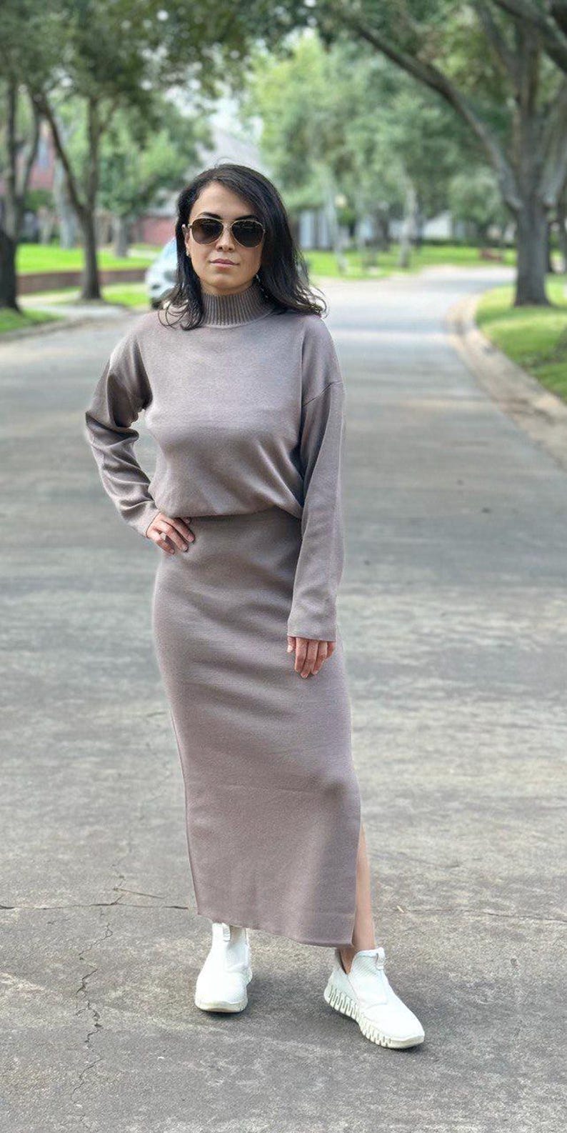 May include: A woman wearing a long-sleeved, light brown sweater dress with a mock neck and a side slit. She is wearing white sneakers and sunglasses.