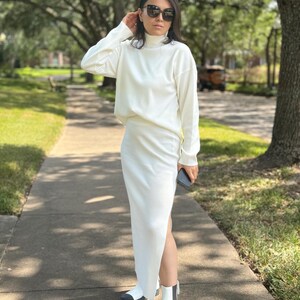 May include: A white turtleneck sweater and matching maxi skirt with a side slit. The outfit is paired with white ankle boots with black accents.