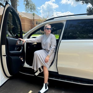May include: A woman wearing a light gray sweater and skirt set, white ankle boots, and sunglasses is getting out of a white SUV.
