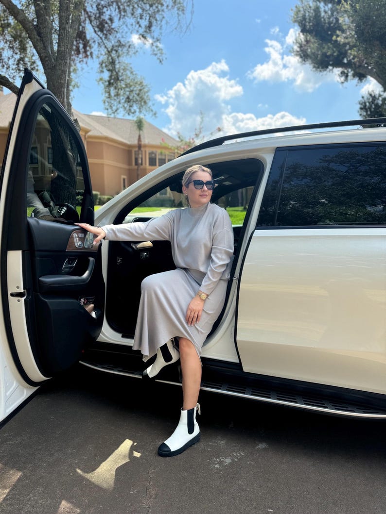 May include: A woman wearing a light gray sweater and skirt set, white ankle boots, and sunglasses is getting out of a white SUV.