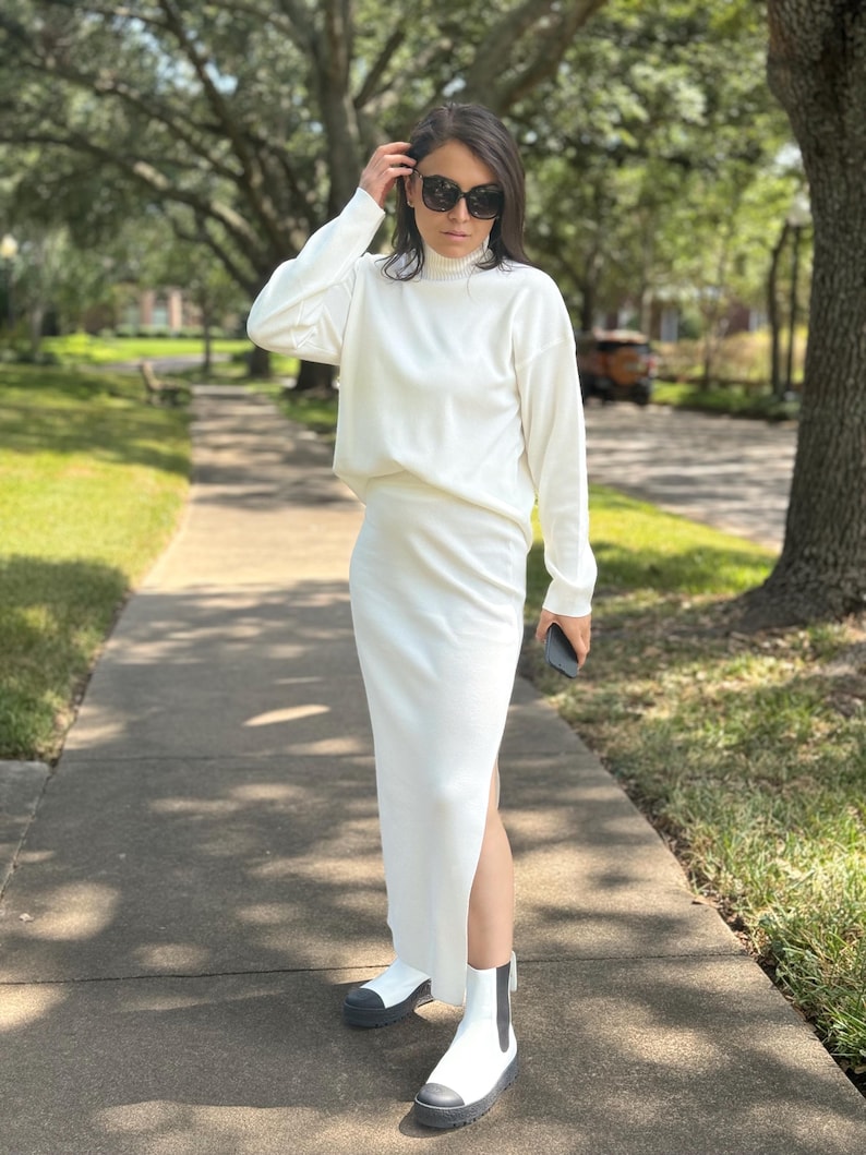 May include: A white knit sweater and matching skirt set with a high neck and a side slit in the skirt. The model is wearing black sunglasses and white ankle boots.