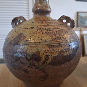 Könnte beinhalten: Eine braune Keramikvase mit rundem Körper und schmalem Hals. Die Vase hat zwei Griffe an den Seiten und eine gesprenkelte Glasur.