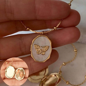 Puede incluir: Un collar de cadena dorada con un colgante de medallón que presenta una mariposa blanca con un strass. El medallón se abre para revelar un espacio para una foto.