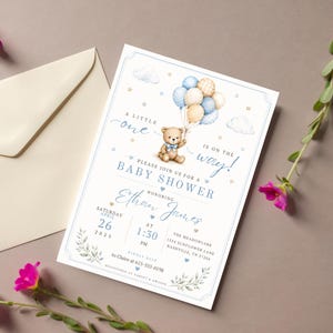 May include: A rectangular baby shower invitation card featuring a watercolor teddy bear holding a cluster of blue, cream, and plaid balloons. The text reads A LITTLE one IS ON THE WAY, PLEASE JOIN US FOR A BABY SHOWER, HONORING Ethan James, SATURDAY APRIL 26 2025 AT 1:30 PM, THE MEADOWLARK 1524 SUNFLOWER LANE NASHVILLE, TN 37204, KINDLY RSVP to Claire at 615-555-0198, REGISTERED AT TARGET & AMAZON.