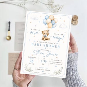 May include: A rectangular baby shower invitation card featuring a watercolor illustration of a teddy bear holding a cluster of blue, cream, and plaid balloons. The text reads A LITTLE one IS ON THE WAY! BABY SHOWER honoring Ethan James.